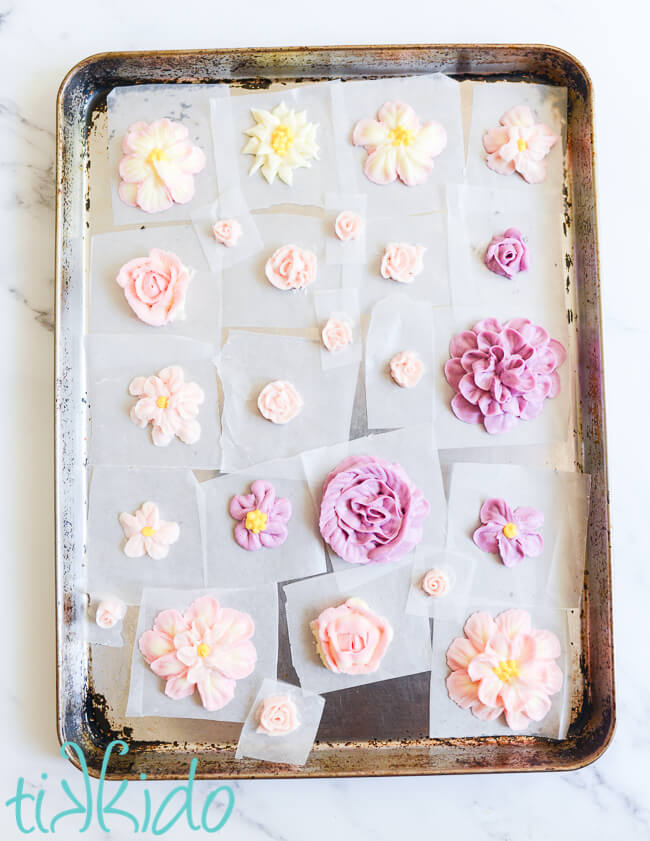 Sheet pan covered with buttercream frosting flowers in various sizes and shapes and shades of pink and purple.  Each flower is on a small square of waxed paper.
