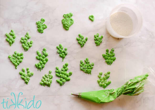 Cactus cupcake toppers piped out of royal icing on waxed paper, with piping bag of green icing and container of white sprinkles