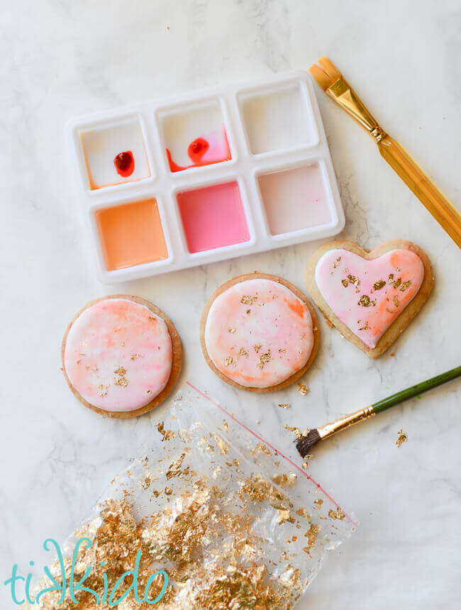 Sugar cookies coated in royal icing, painted with food coloring in tray above the cookies, bag of gold leaf and paint brush below.