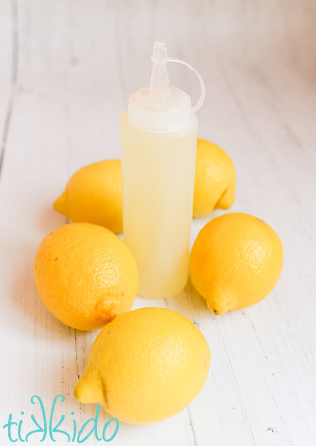 Lemon cake soak in a squeeze bottle, surrounded by five fresh lemons, on a white wooden surface.