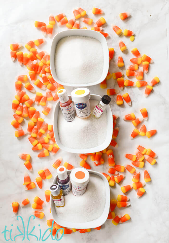 Three bowls of white sugar, with food coloring and flavor containers sitting in the sugar, surrounded by candy corns.