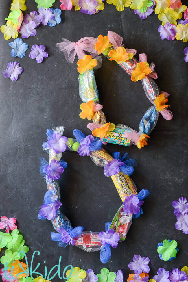 Two candy leis on a black chalkboard background surrounded by colorful silk flowers.