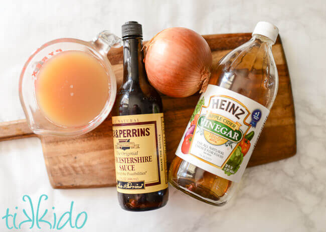 Liquid cooking ingredients for Carolina Pulled Pork, including apple cider, worcestershire sauce, apple cider vinegar, and an onion, displayed on a wooden cutting board.