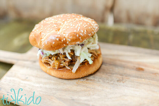 Carolina style pulled pork sandwich topped with coleslaw and on a sesame seed bun, on a wooden cutting board with a black chalkboard backdrop.
