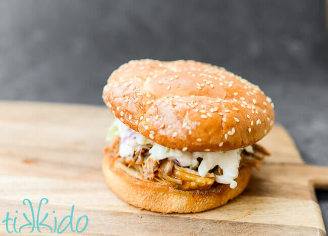 Carolina style pulled pork sandwich topped with coleslaw and on a sesame seed bun, on a wooden cutting board with a black chalkboard backdrop.
