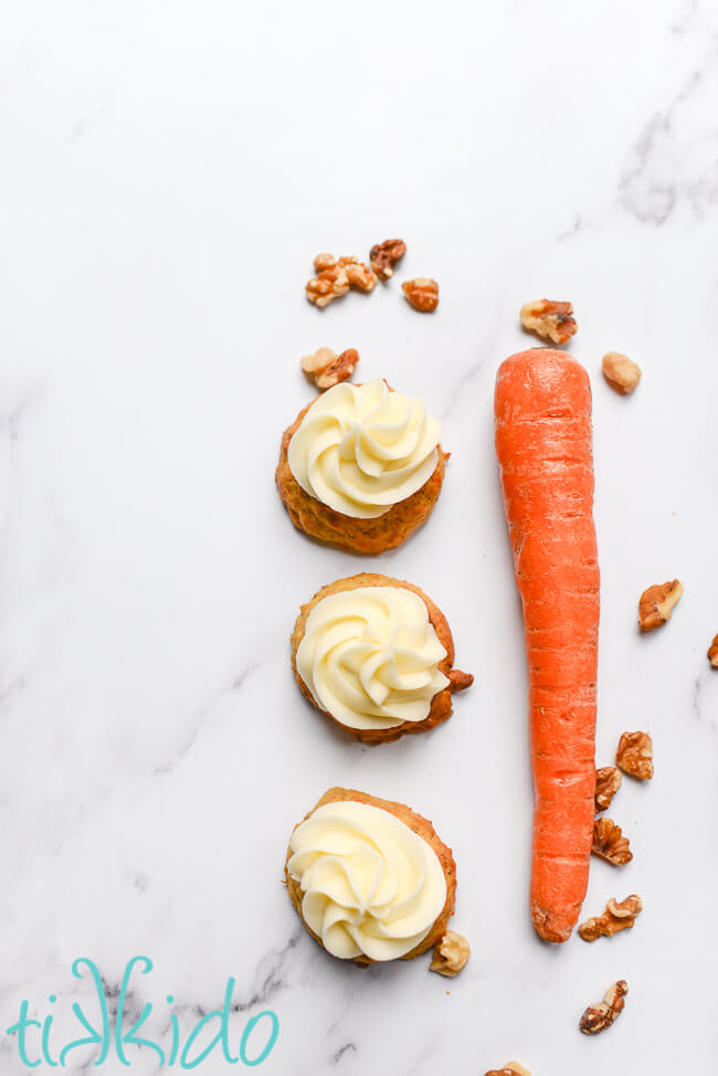 Three carrot cake cookies next to a carrot and chopped walnuts on a white marble surface.