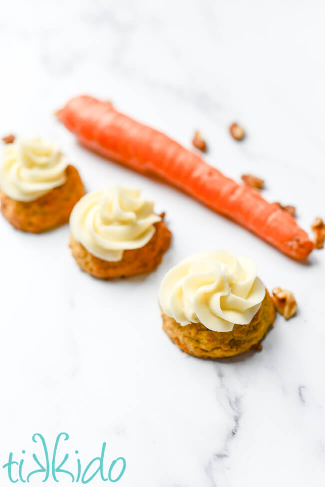 Three carrot cake cookies topped with cream cheese frosting in front of a carrot and walnuts.