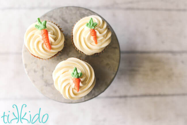 three carrot cake cupcakes with cream cheese icing and an edible chocolate carrot cupcake topper on a galvanized metal cake stand.