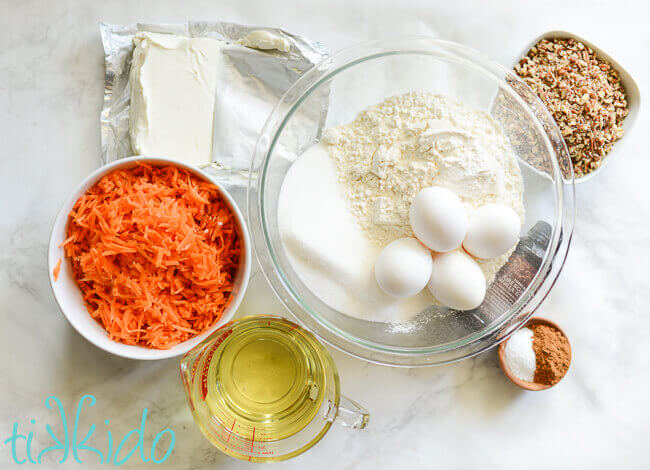 Ingredients for carrot cake, including carrots, oil, cream cheese, flour, sugar, eggs, baking powder, cinnamon, salt, vanilla, and chopped nuts) arranged on a white marble background.