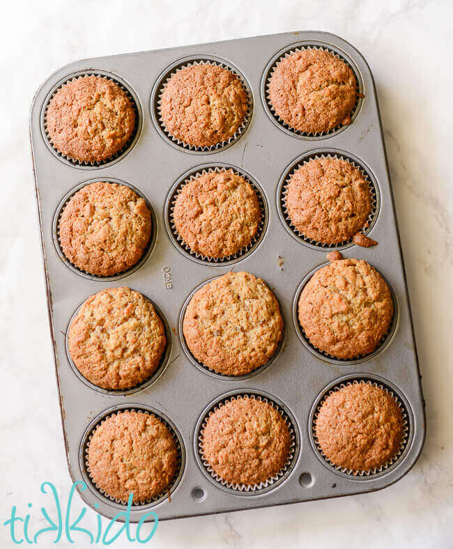 A dozen baked carrot cake cupcakes in a silver muffin tin on a white marble background