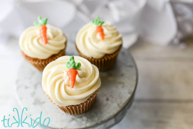 Three carrot cake cupcakes swirled with cream cheese icing and topped with a small chocolate edible carrot cupcake topper.  The cupcakes sit on a galvanized metal cake stand on a weathered white wood backdrop.