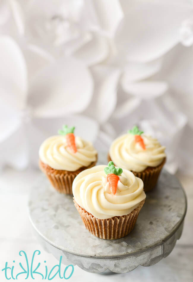 three carrot cake cupcakes with cream cheese icing and an edible chocolate carrot cupcake topper on a galvanized metal cake stand.