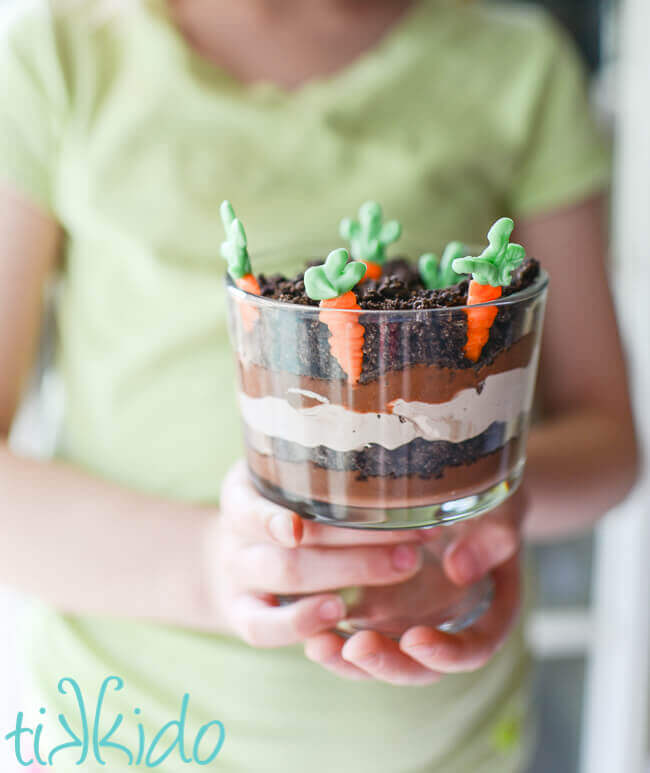 miniature trifle dishe filled with layers of chocolate pudding, chocolate whipped cream, and crushed oreo dirt, with white chocolate carrots planted in the top layer. Held by a girl wearing a light green shirt.