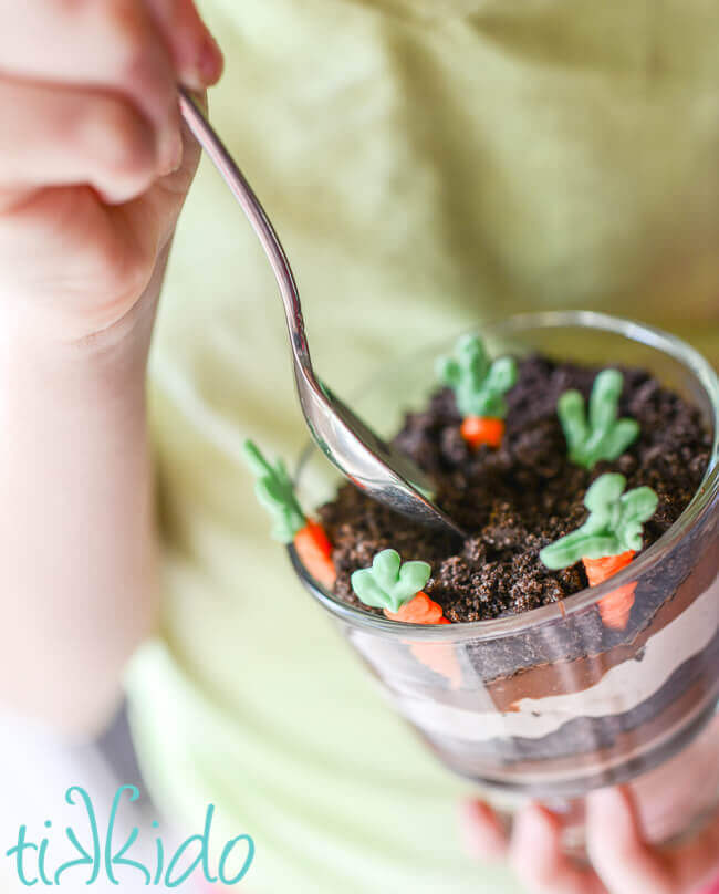 miniature trifle dishe filled with layers of chocolate pudding, chocolate whipped cream, and crushed oreo dirt, with white chocolate carrots planted in the top layer. Held by a girl wearing a light green shirt.