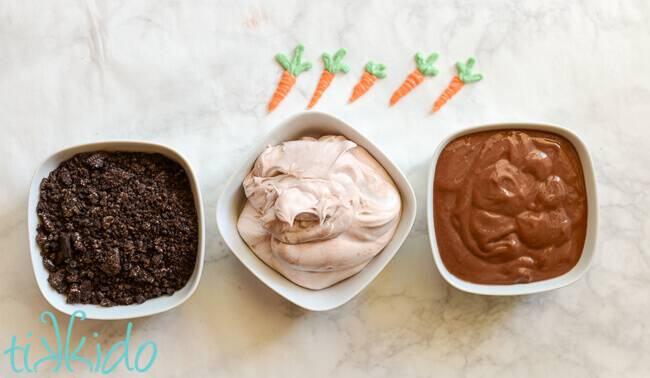 Bowls of crushed oreo dirt, Chocolate whipped topping, and chocolate pudding.  Five small chocolate carrots arranged above the dishes, on a white marble background.
