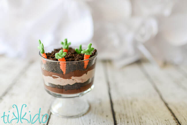 miniature trifle dishe filled with layers of chocolate pudding, chocolate whipped cream, and crushed oreo dirt, with white chocolate carrots planted in the top layer.