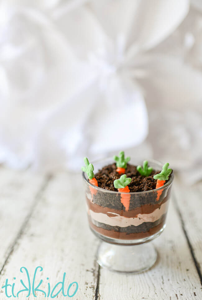 Two miniature trifle dishes filled with layers of chocolate pudding, chocolate whipped cream, and crushed oreo dirt, with white chocolate carrots planted in the top layer.