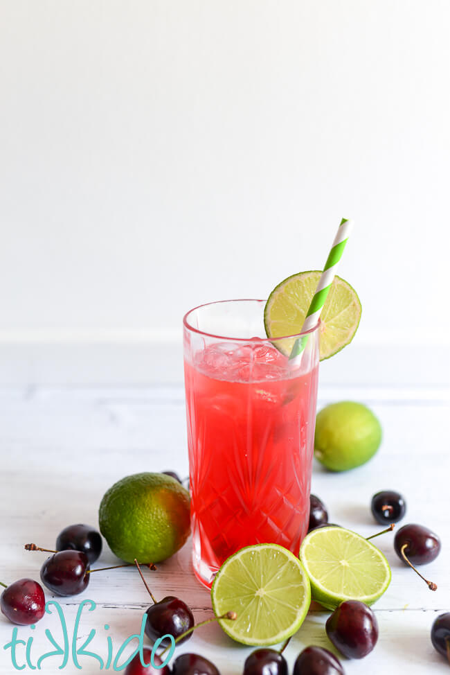 Tall crystal glass full of all-natural cherry limeade, garnished with a round slice of lime and a green and white striped paper straw.  Fresh cherries and limes surround the glass of Cherry Limeade on a white wooden surface.