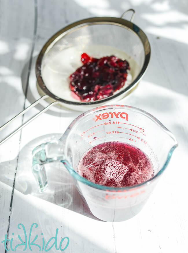 Pyrex measuring cup full of cherry simple syrup, with cherries in a fine mesh strainer resting behind the measuring cup.