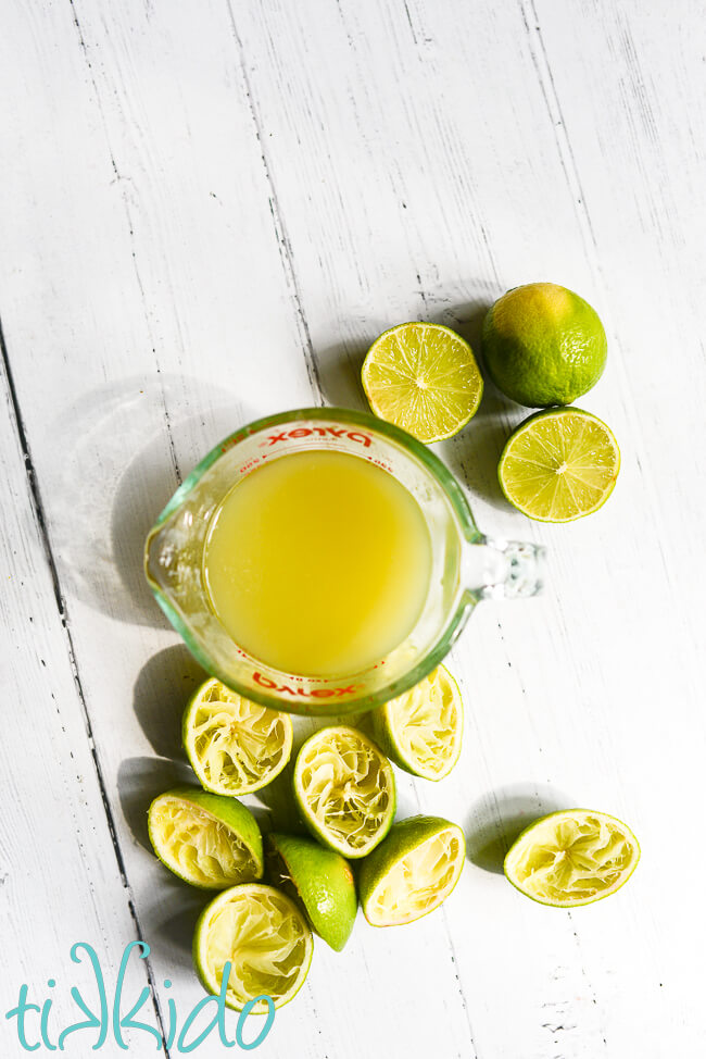 A pyrex measuring cup full of freshly squeezed lime juice, surrounded by whole limes and lime halves that have already been juiced, on a white wooden surface.