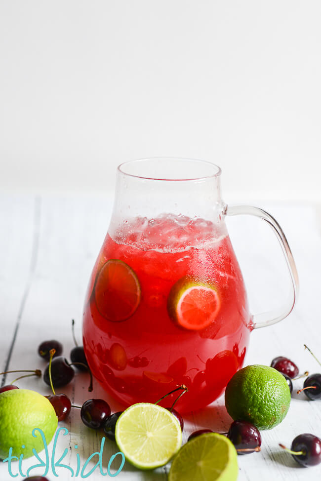 Pitcher of from scratch Cherry Limeade surrounded by fresh limes and cherries, on a white wooden surface.