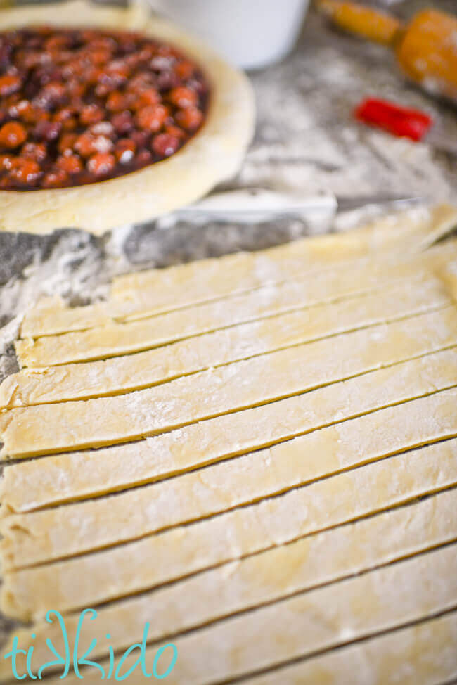 Pie crust rolled out and cut into strips with a pie tin filled with cherry pie filling in the background.