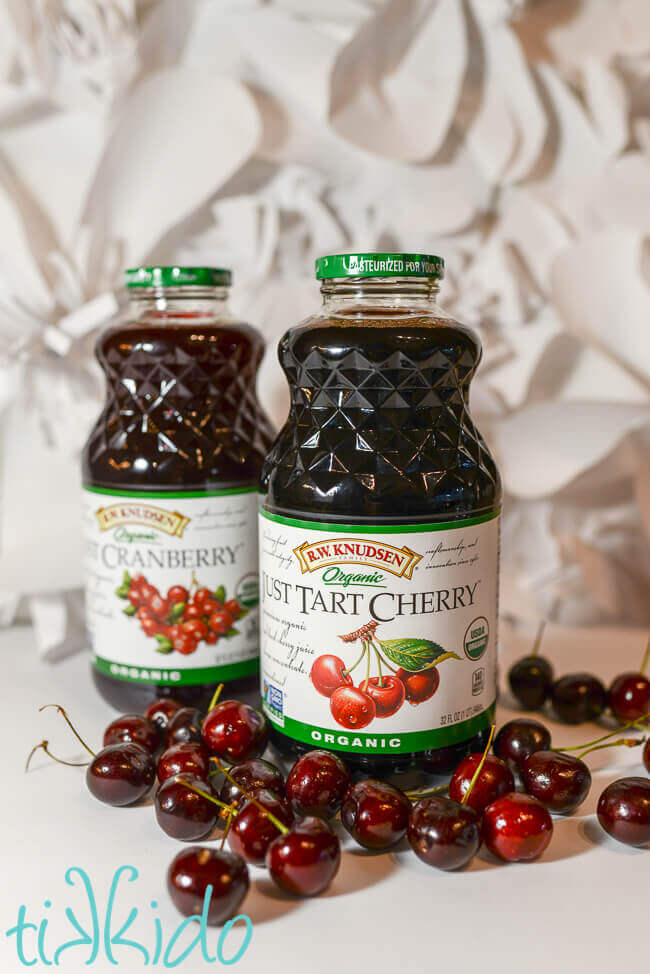 A jar of tart cherry juice and a a jar of cranberry juice surrounded by fresh cherries.