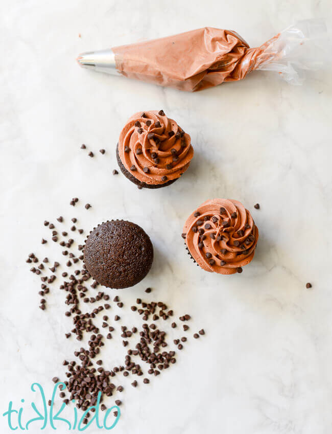 Three chocolate cupcakes made with Crazy Cake Recipe being topped with chocolate icing and mini chocolate chips on a white marble background.