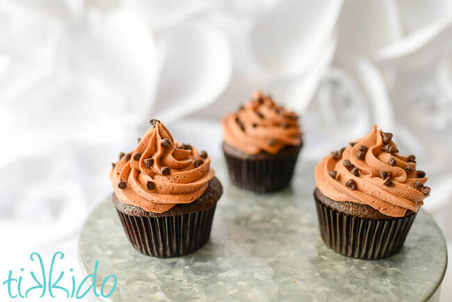 Three chocolate Crazy Cake cupcakes topped with chocolate icing and mini chocolate chips on a metal cake stand on a white background.