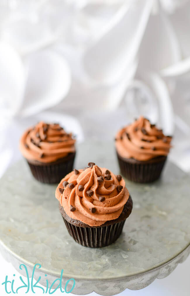 Three chocolate cupcakes topped with chocolate icing and mini chocolate chips on a metal cake stand on a white background.