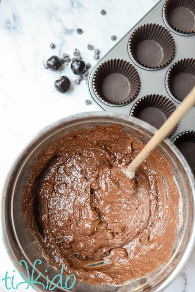 Batter for chocolate cherry muffins in a silver mixing bowl next to an empty muffin tin.