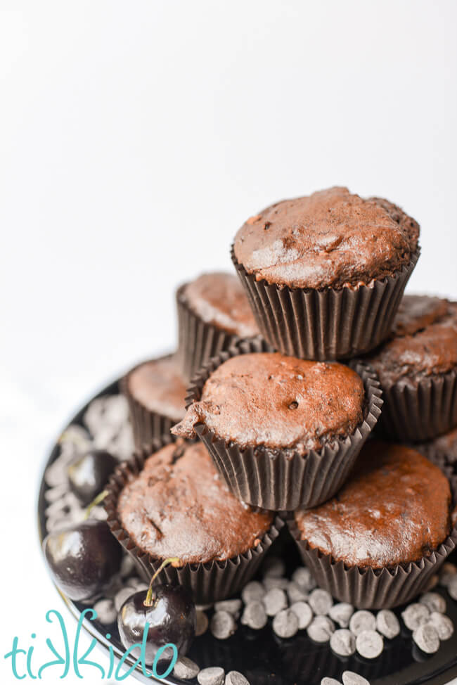 chocolate cherry muffins stacked on a black cake plate, surrounded by fresh cherries and chocolate chips.