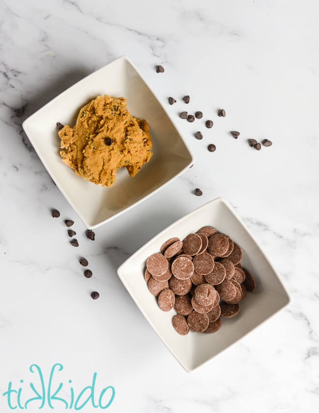 Bowl of edible chocolate chip cookie dough and a bowl of chocolate melts on a white marble surface, with chocolate chips scattered around.