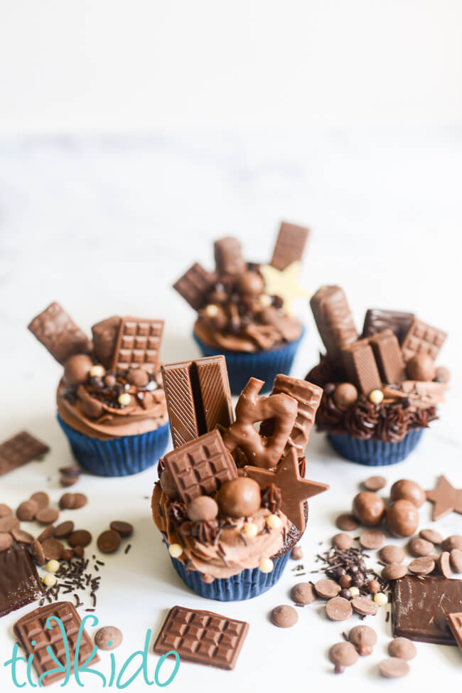 Four chocolate explosion cupcakes on a white marble surface, with chocolate candies and sprinkles scattered around the cupcakes.