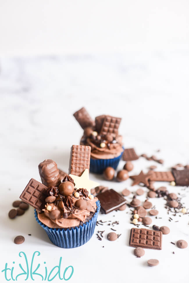 Two chocolate explosion cupcakes surrounded by scattered chocolate candies and chocolate sprinkles on a white marble surface.