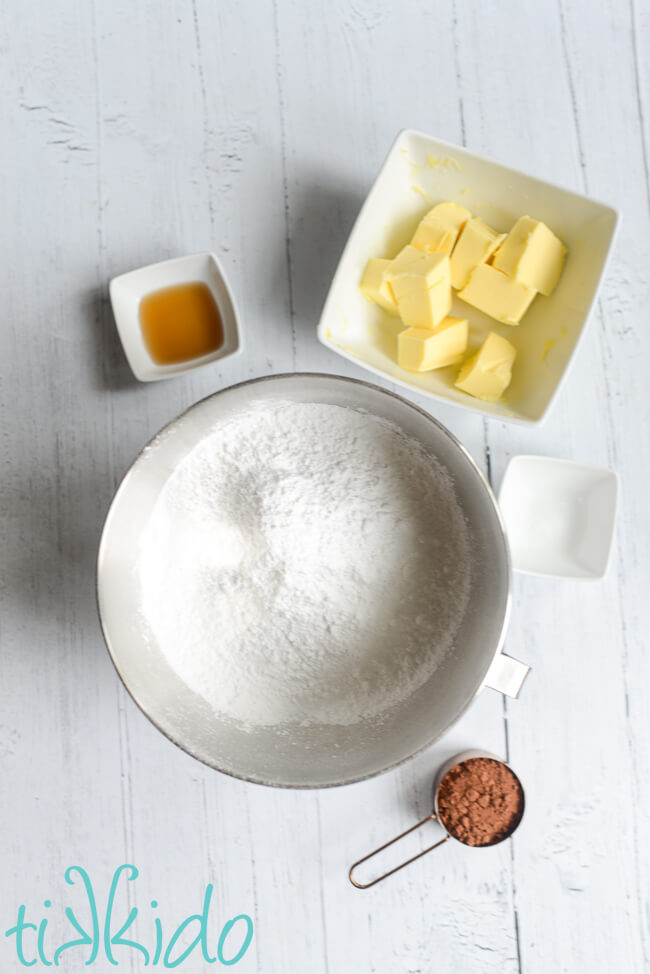 Ingredients for chocolate icing with cocoa powder measured out in bowls on a white wooden surface.