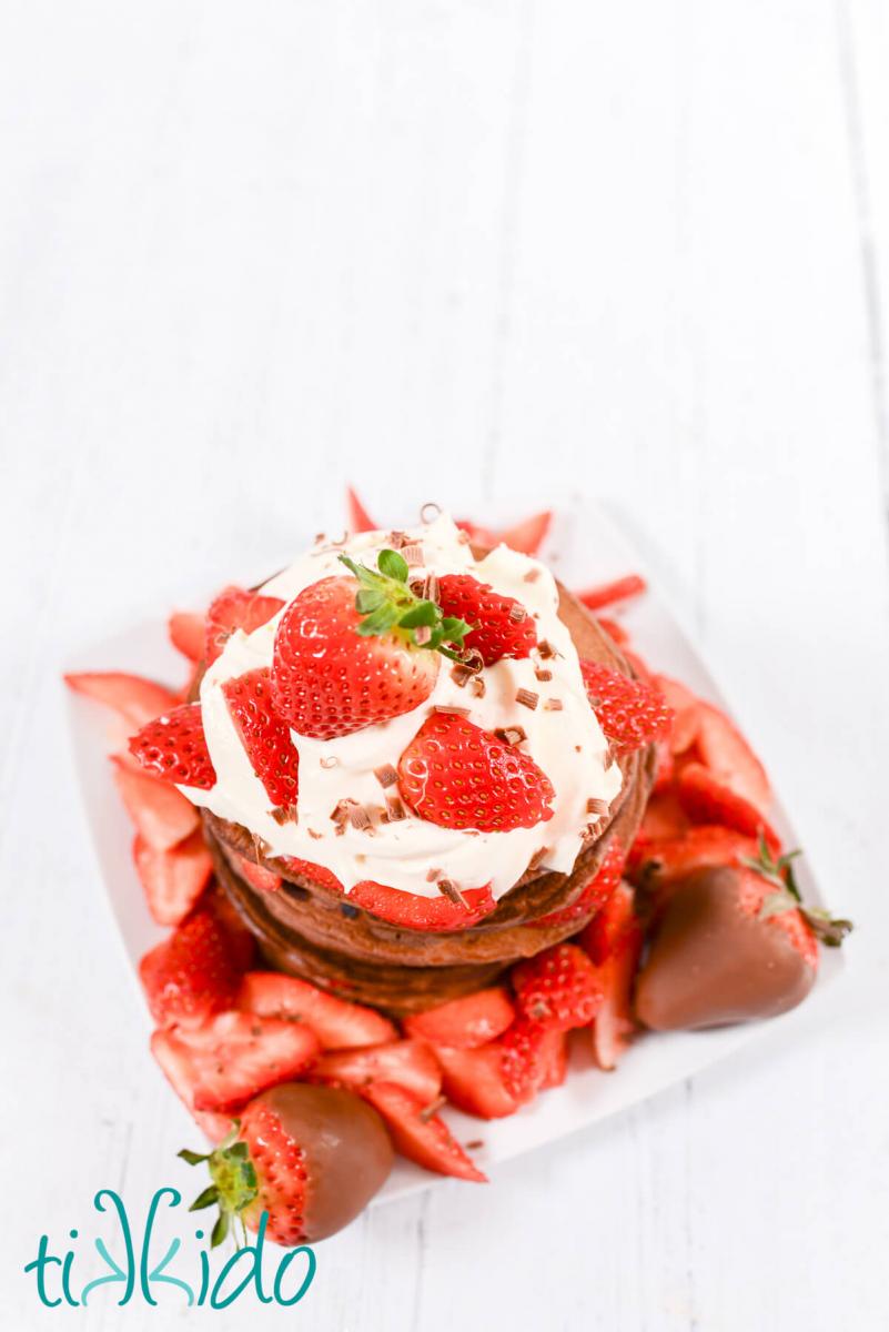 Top down view of a tall stack of chocolate pancakes topped with whipped cream and fresh strawberries.