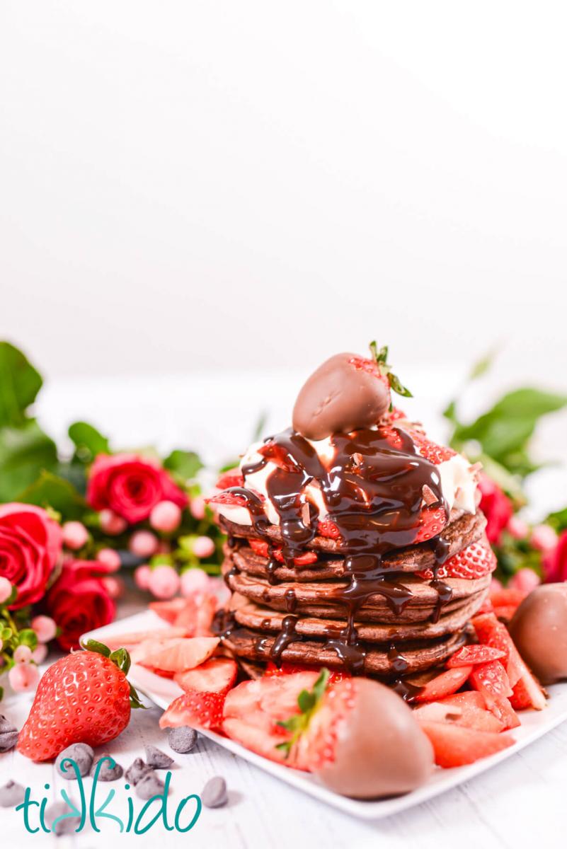 Tall stack of chocolate pancakes topped with whipped cream, fresh strawberry slices, chocolate syrup, and a chocolate covered strawberry.
