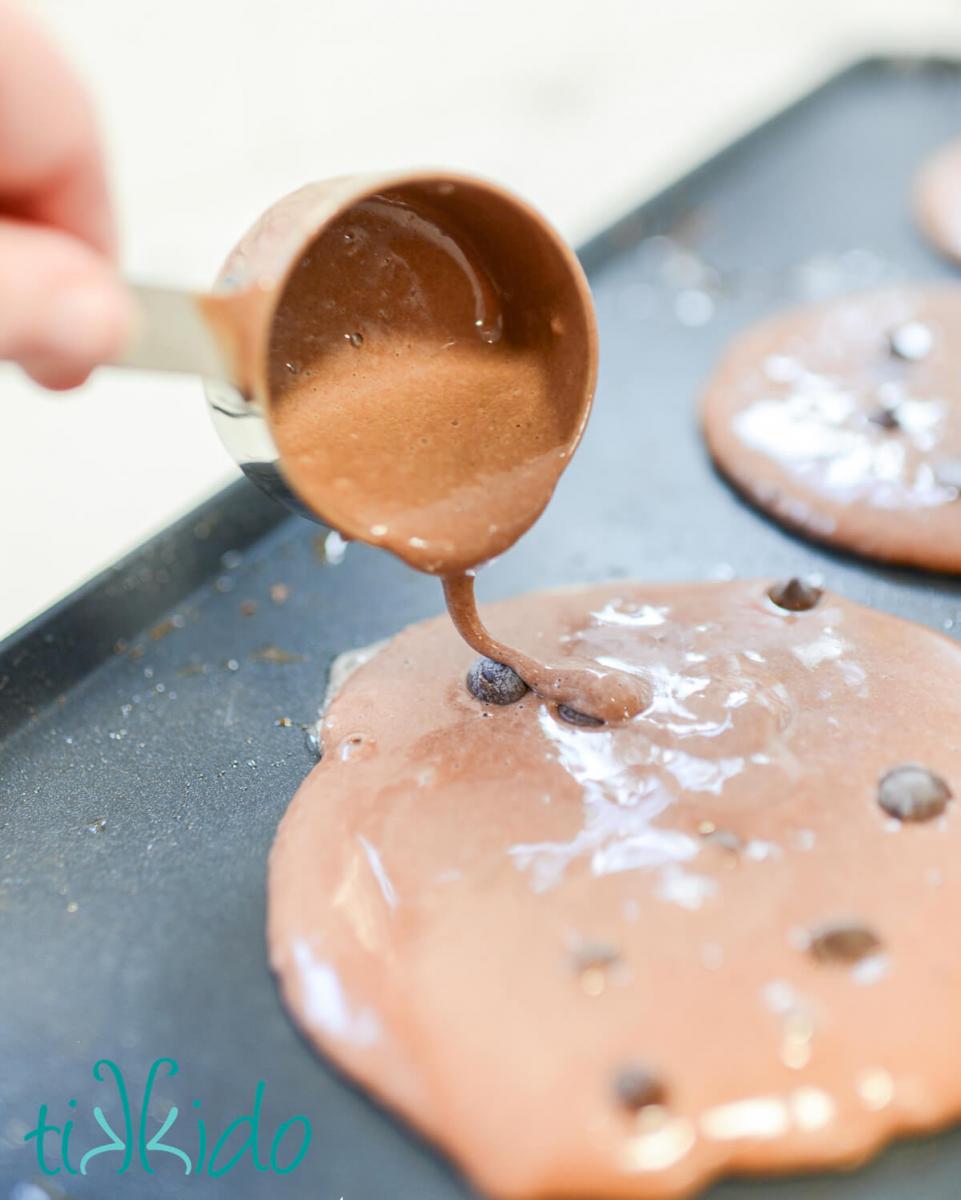 Chocolate pancakes with chocolate chips being cooked on an electric griddle.  The chocolate chips are being covered with extra chocolate pancake batter.