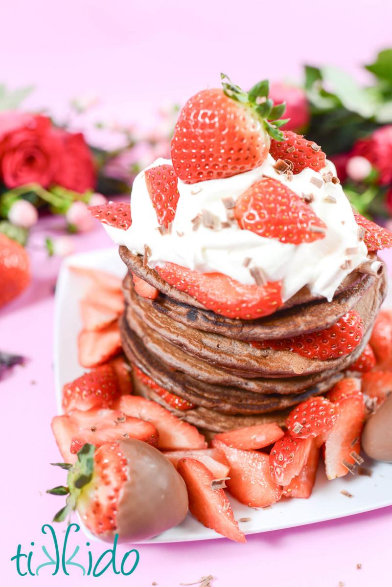 Stack of chocolate pancakes topped with a dollop of whipped cream, covered in sliced fresh strawberries, and chocolate curls.  The plate sits on a pink surface, and roses and fresh strawberries are in the background of the photo.