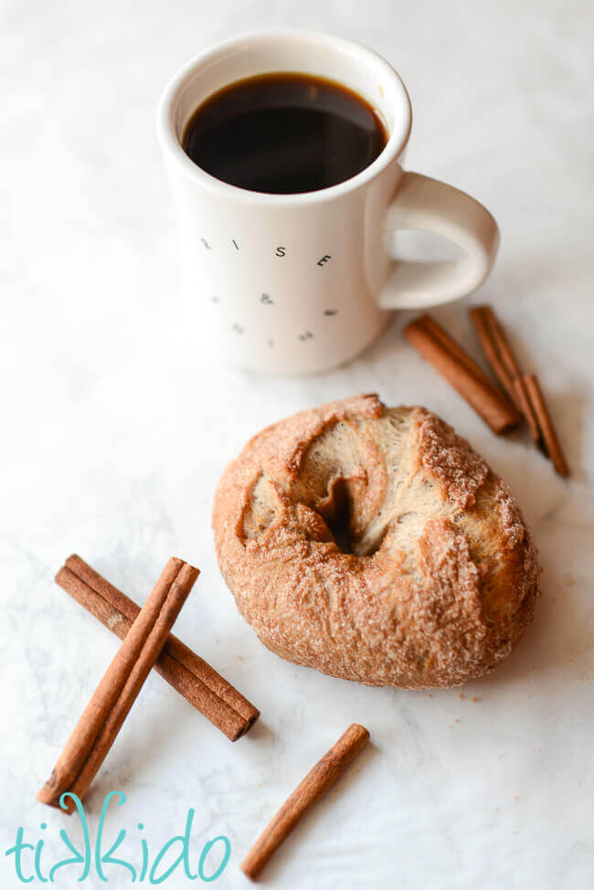 Cinnamon sugar bagels on a white surface surrounded by cinnamon sticks and a cup of coffee.