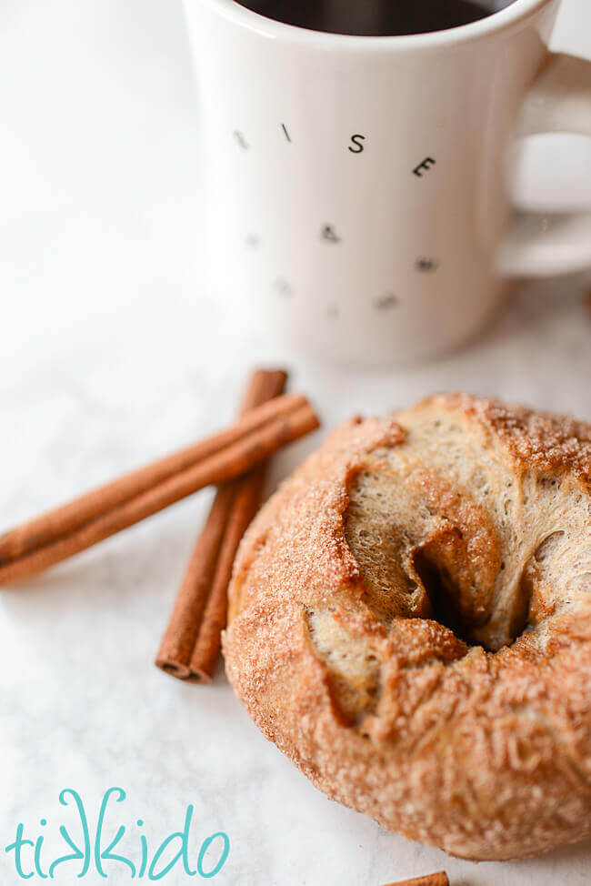 Cinnamon sugar bagels on a white surface surrounded by cinnamon sticks and a cup of coffee.