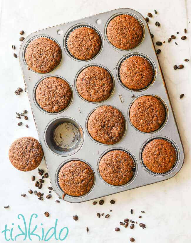 Coffee cupcakes in a cupcake tin, surrounded by coffee beans on a white marble surface.