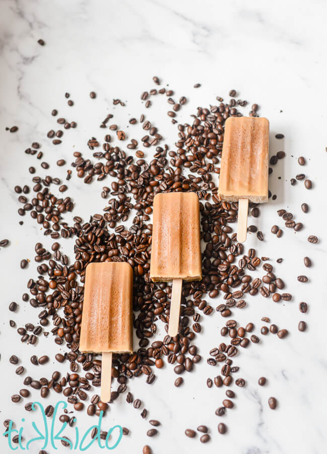 Overhead view of three coffee popsicles sitting on a bed of whole coffee beans on a white marble surface.