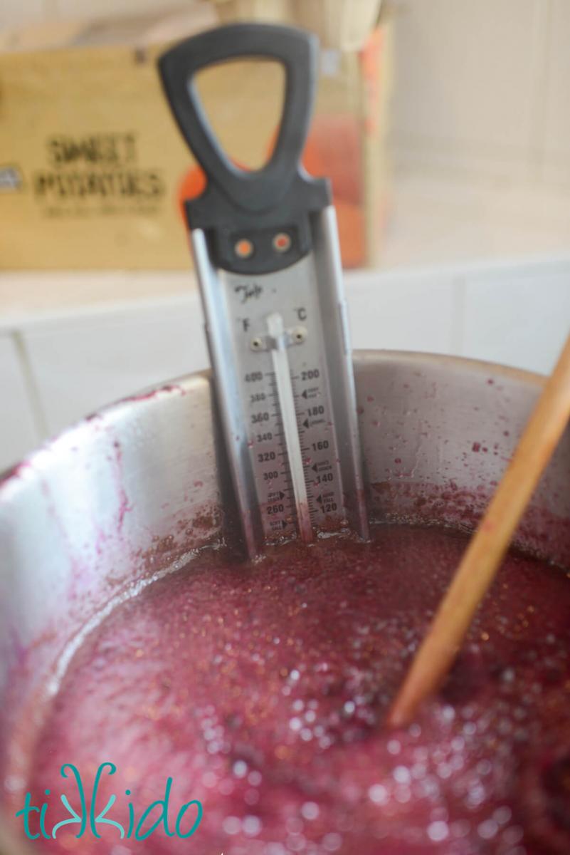 Concord grape jam being cooked in a stock pot, stirred with a wooden spoon, with a candy thermometer clipped to the pot.