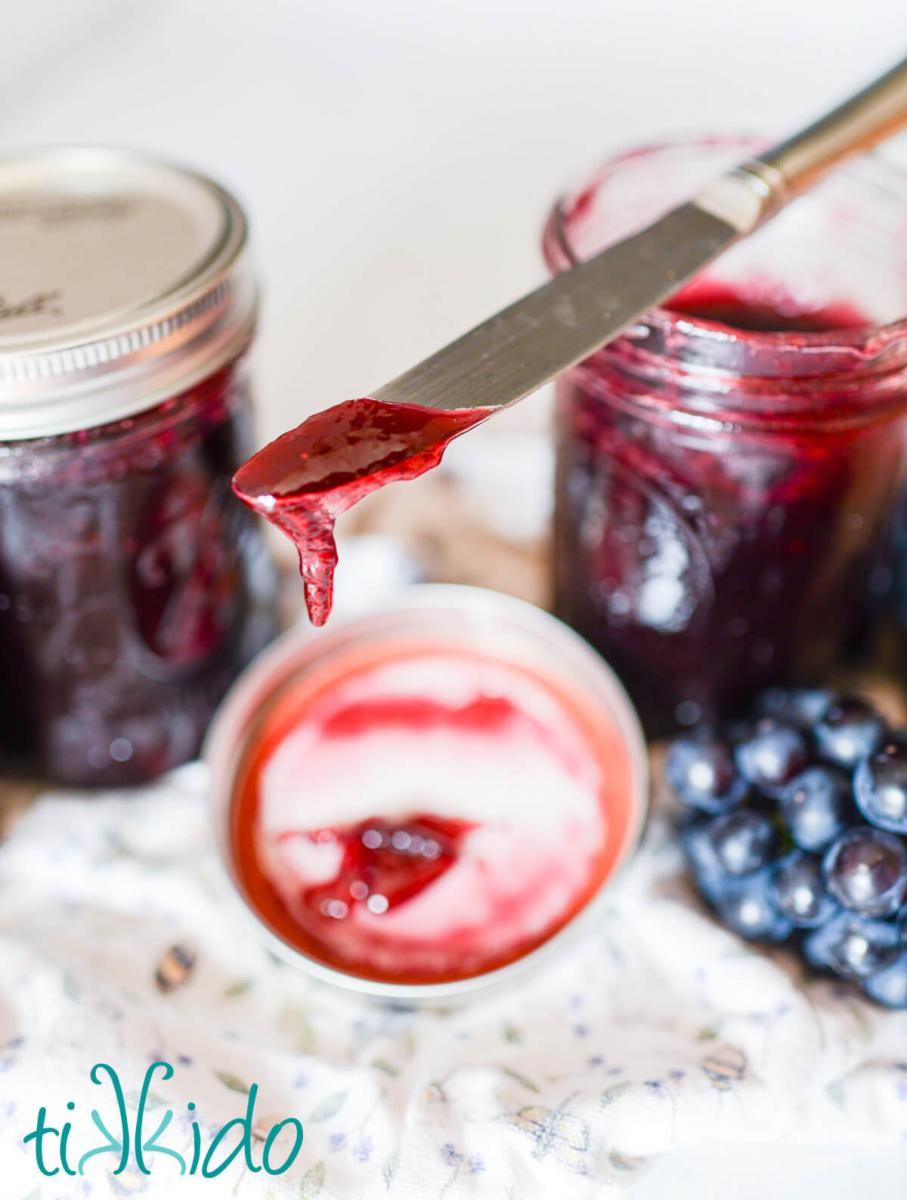 Grape jam dripping off the end of a knife onto the jam lid below.  The knife rests on the open jar of concord grape jam.