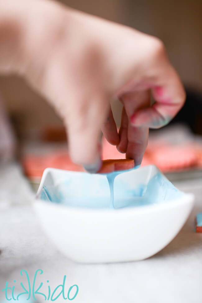 Heart shaped cookie being dipped in thin blue royal icing to make Conversation Heart Cookies