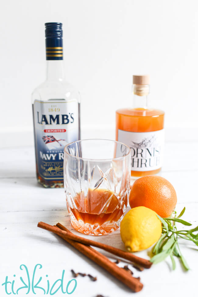 Rum and Shrub drink from Cornwall surrounded by a bottle of rum, a bottle of Cornish shrub, and fresh citrus, herbs, and spices.