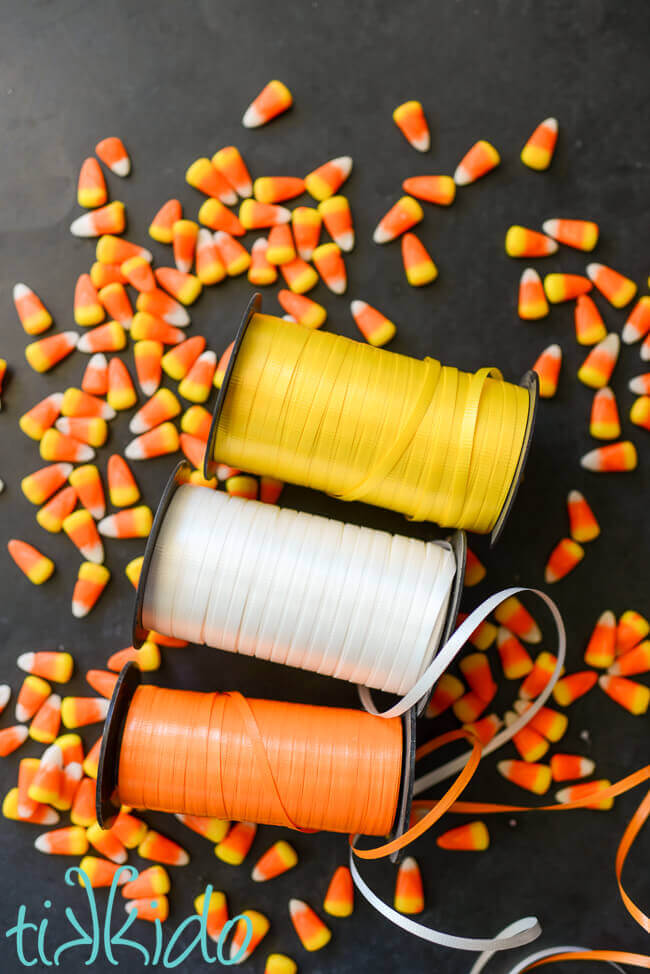 Yellow, white, and orange spools of curling ribbon on a black chalkboard background, surrounded by candy corn candies.