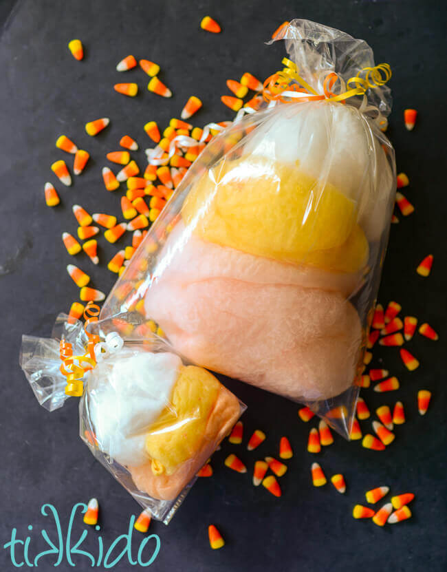 Two bags of white, yellow, and orange cotton candy surrounded by candy corn candies on a black background.